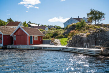 Rød sjøbod og tilhørende brygge ved vannkanten. Nydelige omgivelser med hus på høydene og sol som reflekteres i vannet.