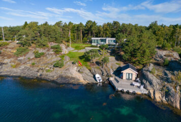 Dronebilde av hyttetomt med hytte og naust ved kysten. Omgitt av natur, med klipper, skog og utsikt over havet.